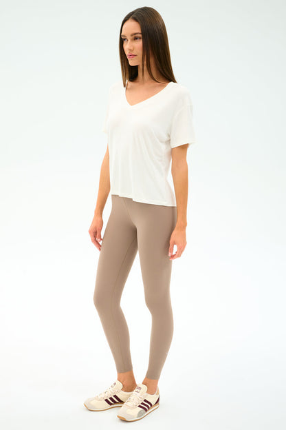 A woman stands against a plain background wearing the SPLITS59 Georgia Jersey V-Neck in white, paired with beige leggings and beige sneakers featuring brown stripes.