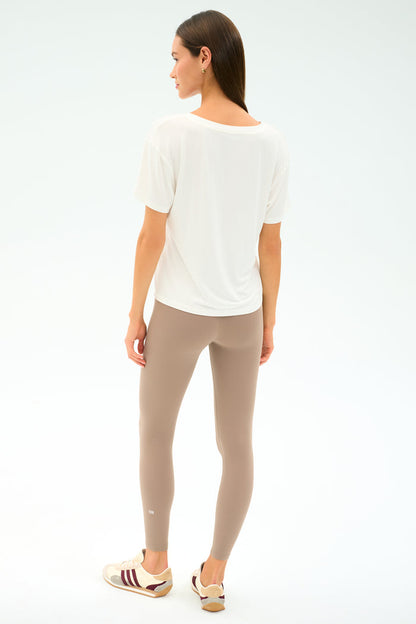 A woman faces away, wearing the SPLITS59 Georgia Jersey V-Neck in white and taupe spandex-blend leggings, paired with beige sneakers featuring maroon stripes, set against a plain light background.