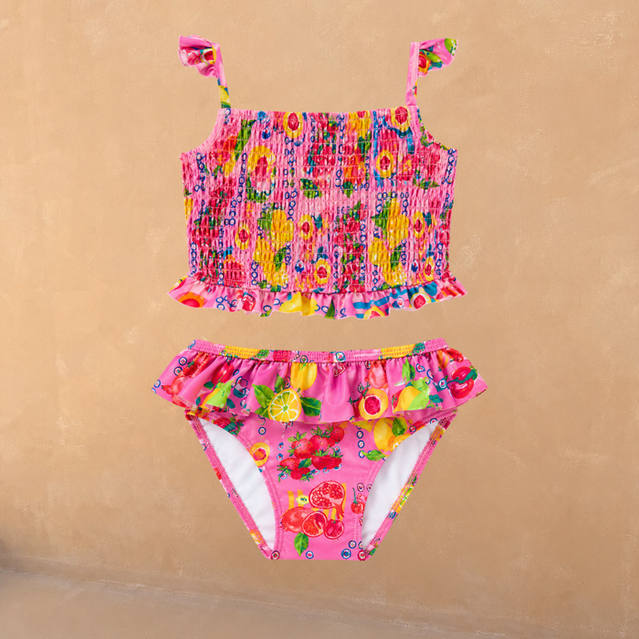 Girls pink Mediterranean fruit print bikini set with a shirred top and frill bottoms on a neutral background.