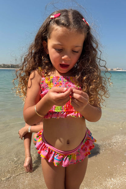 Girls Pink Fruit Crop Bikini Set with a Mediterranean fruit print, shirred bandeau top and frill bottoms at the beach.
