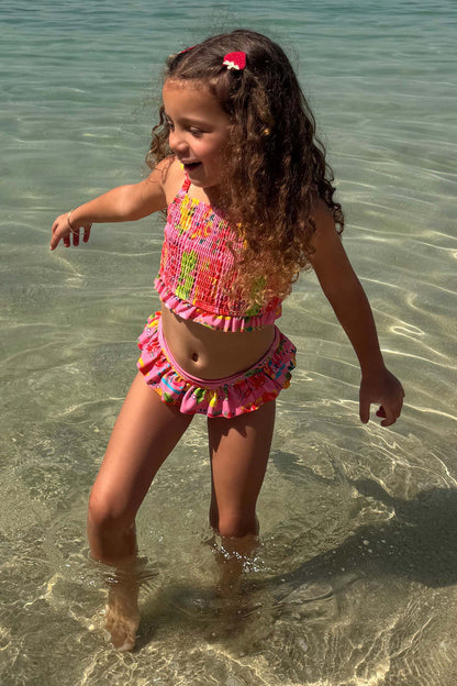 Pink Fruit Crop Bikini Set with a fruit print, shirred top and frill bottoms on a young girl in the sea.