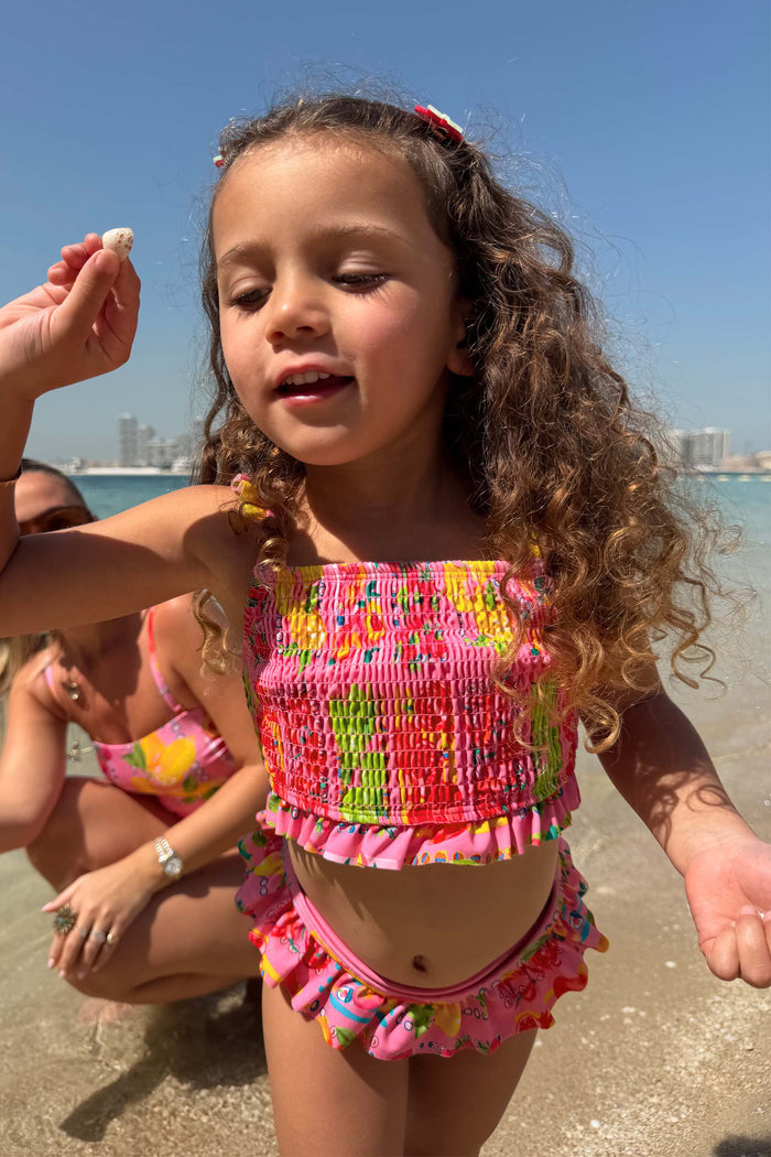 Girls Pink Fruit Crop Bikini Set with a shirred bandeau top and frill bottoms on a girl at the beach.