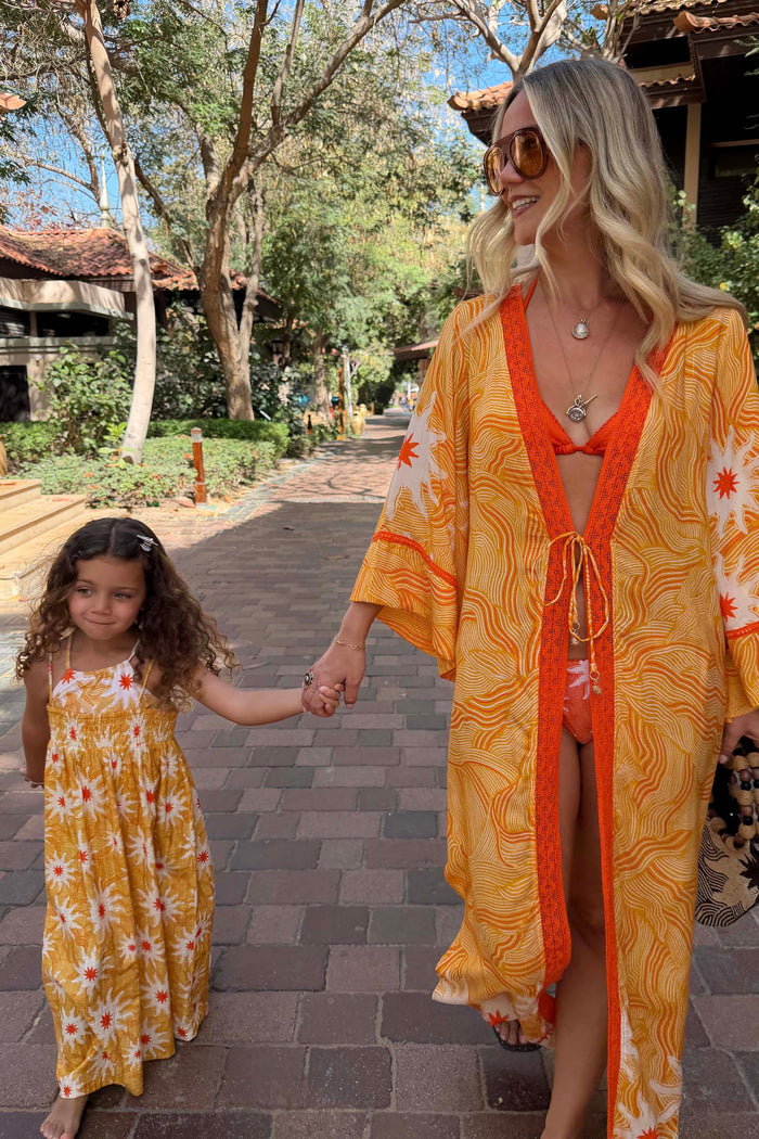 Girl's yellow cotton maxi sun dress with an orange sun print and shirred top, walking with a matching adult on vacation.