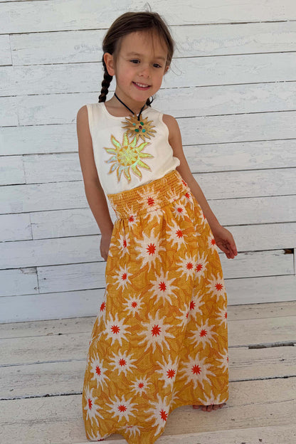 Yellow cotton maxi dress with an orange sun print and shirred waist on a girl against a white wooden wall.