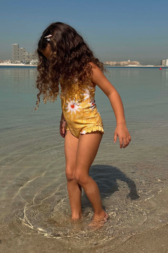 Girl's Yellow Sun Swimsuit with a playful sun print and soft frill trims on a girl standing in shallow water.