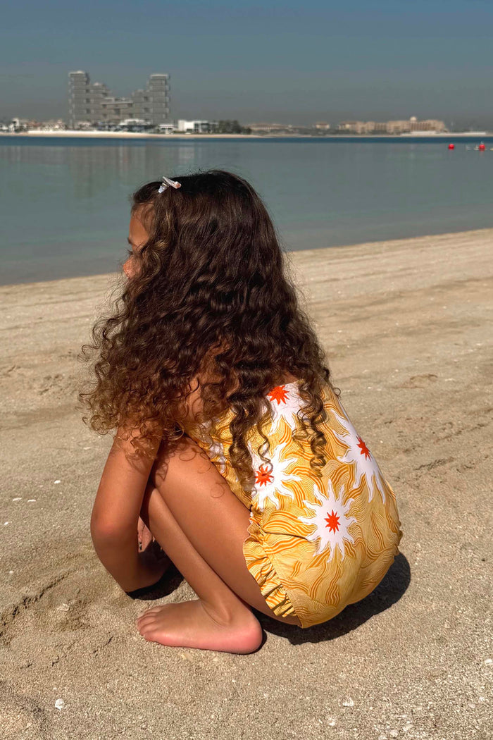Girl's Yellow Sun Swimsuit with sun print and frill detailing on a young girl crouching on a beach.