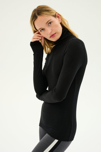 A person with long hair wearing a black Jackson Rib Full Length Turtleneck by SPLITS59 and gray leggings stands against a plain background.