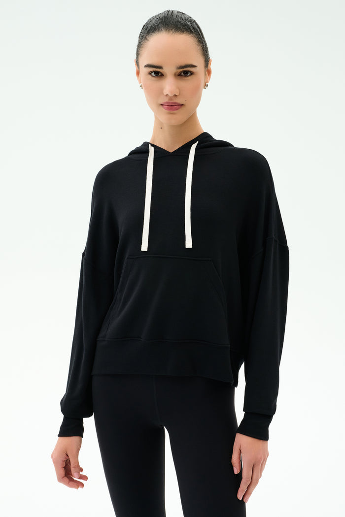 A person wears the SPLITS59 Joey Fleece Hoodie in black with white drawstrings and black spandex-blend leggings, standing against a plain white background.