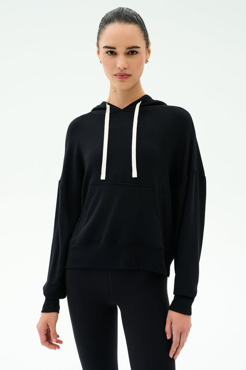 A person wears the SPLITS59 Joey Fleece Hoodie in black with white drawstrings and black spandex-blend leggings, standing against a plain white background.