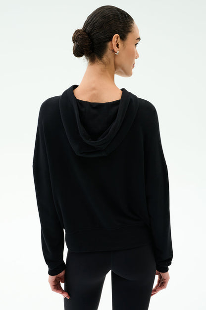A woman with dark hair in a low bun is seen from behind wearing the SPLITS59 Joey Fleece Hoodie in black against a plain light background.