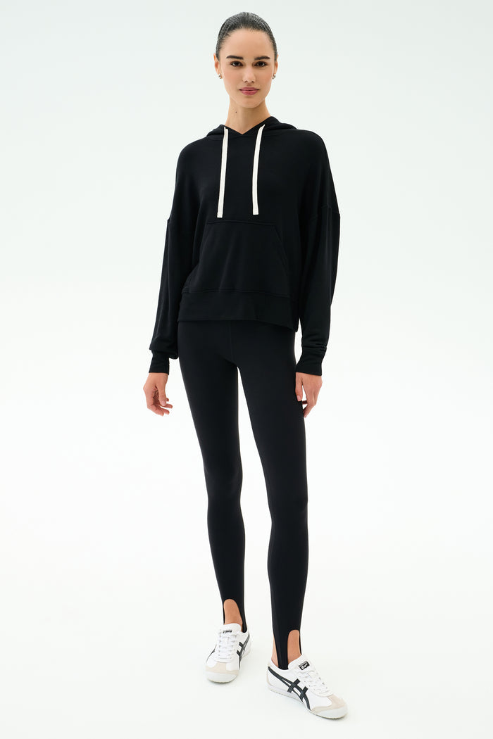 A person stands facing forward in a SPLITS59 Joey Fleece Hoodie in black, black spandex blend leggings with stirrups, and white sneakers against a plain white background.