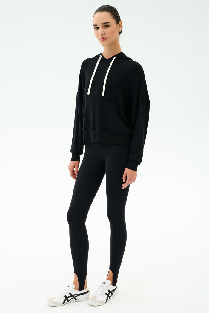 A person stands facing forward against a plain white background, wearing the SPLITS59 Joey Fleece Hoodie in black, black leggings, and white sneakers with black accents.
