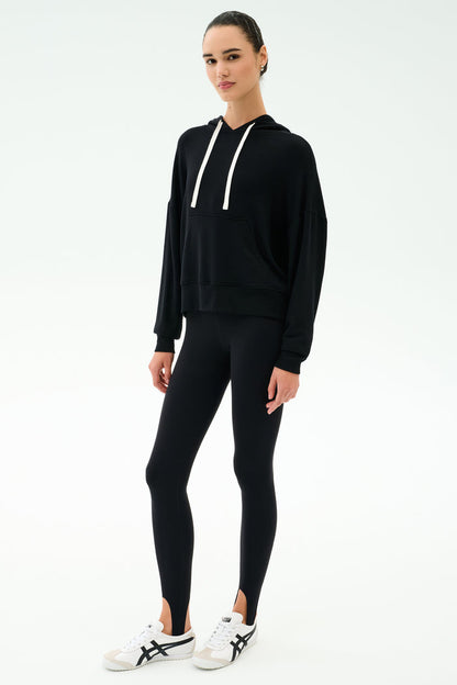 A person stands facing forward against a plain white background, wearing the SPLITS59 Joey Fleece Hoodie in black, black leggings, and white sneakers with black accents.