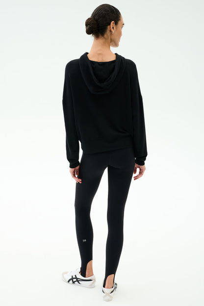 A woman stands with her back to the camera, wearing the SPLITS59 Joey Fleece Hoodie in black and matching leggings, paired with white sneakers, against a plain white background.