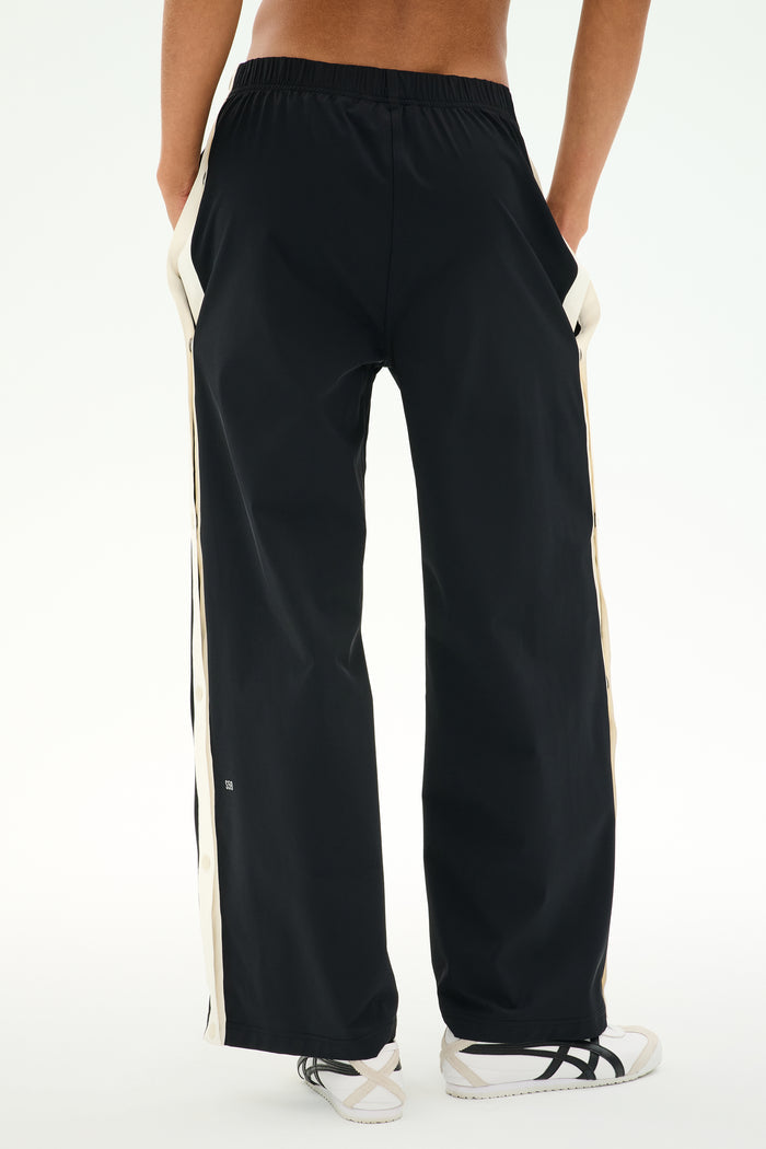 A person is seen from behind with hands in pockets, wearing SPLITS59's Jordan Nylon Twill Tearaway pants in Black/Creme, featuring white side stripes and crafted from a comfortable blend of nylon and spandex.