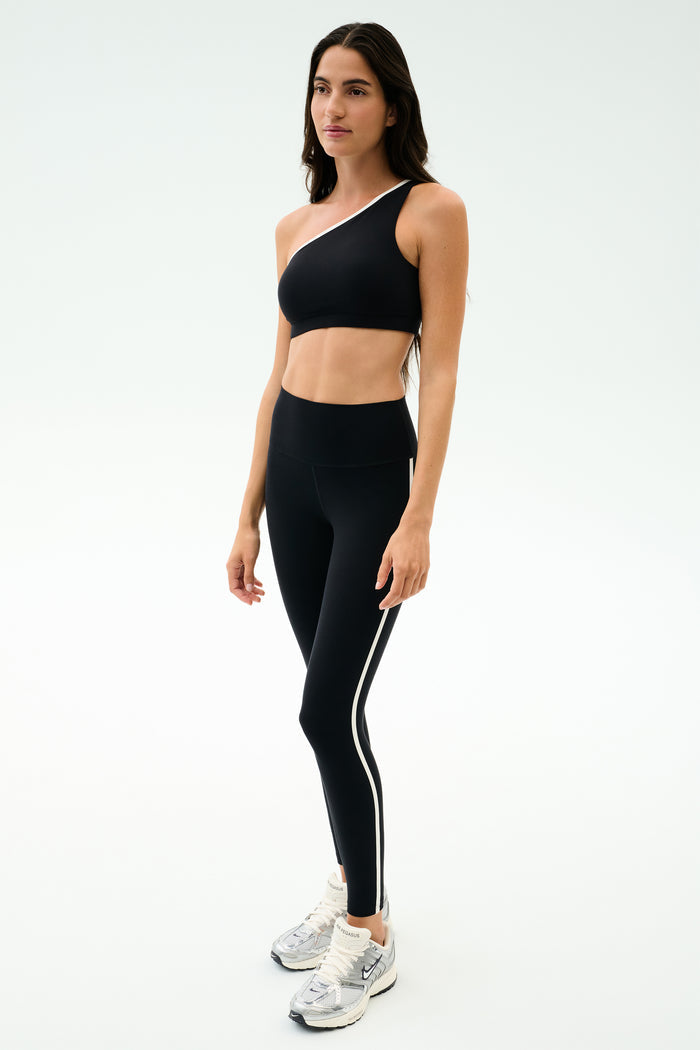 A woman wears the SPLITS59 Josie Airweight One Shoulder Bra in Black/White with black leggings featuring a white side stripe and white athletic shoes, standing against a plain light background—ideal for hot yoga sessions.