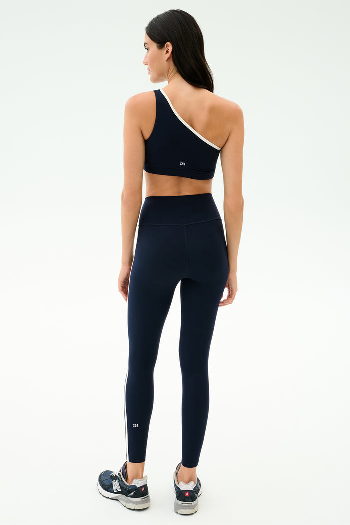 A woman faces away, wearing the SPLITS59 Amber Airweight High Waist 7/8 leggings in Indigo/White with a matching one-shoulder sports bra—perfect for hot yoga—paired with black sneakers against a plain white background.