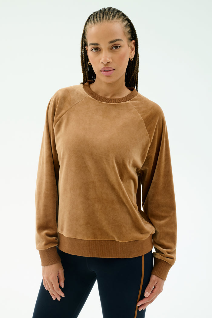 A woman with braided hair wears the SPLITS59 Joy Velour Crewneck in Honey, paired with dark side-striped stretch pants, standing against a plain light background.