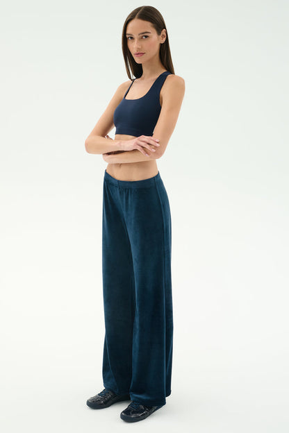 A woman stands with arms crossed, wearing the SPLITS59 Joy Velour Pant in Indigo and a navy crop top, paired with black shoes against a plain light background.