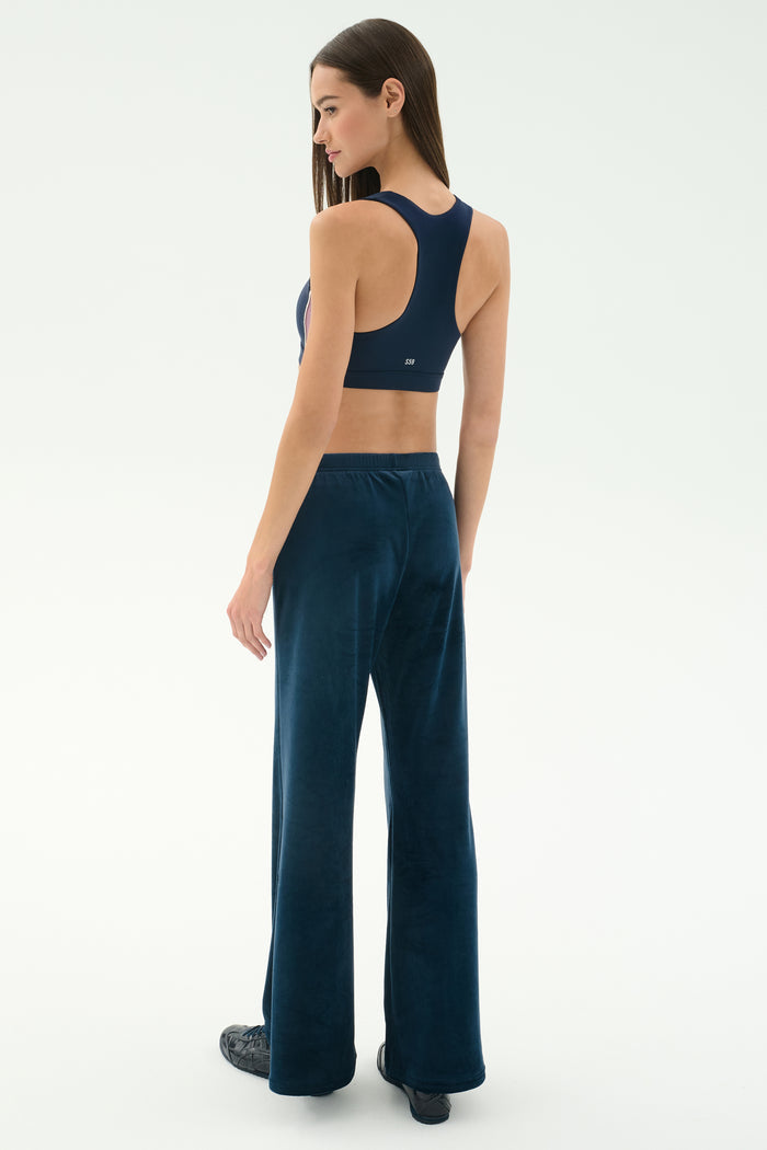 A woman faces away in SPLITS59 Joy Velour Pant in Indigo and a navy sports bra with black shoes, set against a plain light background.