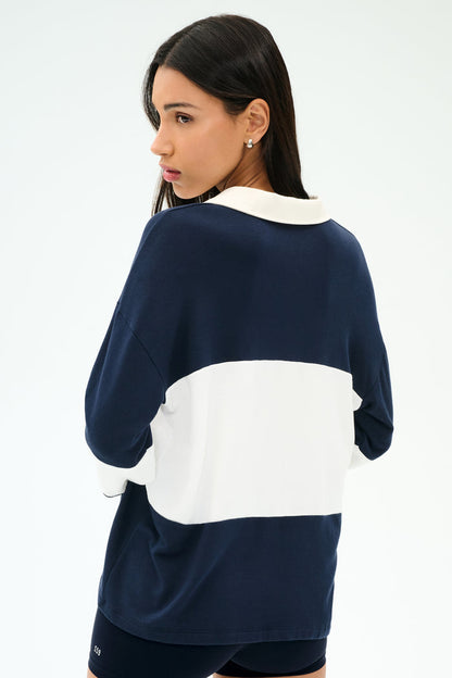 A woman with long dark hair models the SPLITS59 Katie Fleece Rugby in Indigo/White, glancing over her shoulder against a plain light background.