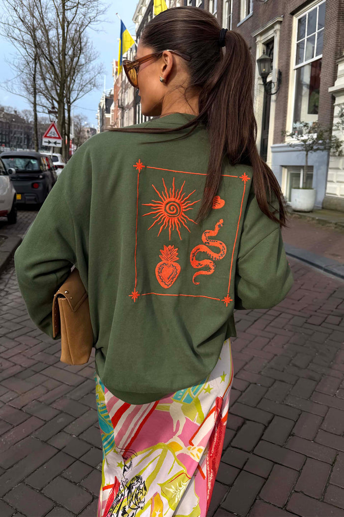 Khaki sweatshirt with orange sun and snake embroidery on the back, styled for a casual London look.
