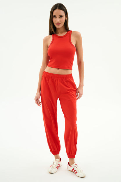 A woman stands against a plain background wearing the SPLITS59 Kiki Rib Crop Tank in Lotus Red, paired with red jogger pants and white sneakers with red stripes—ideal for gym sessions or casual outings.