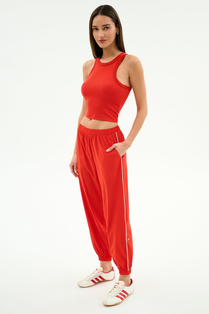 Woman wearing the SPLITS59 Kiki Rib Crop Tank in Lotus Red, paired with red joggers and white Adidas sneakers, standing against a white background. A chic sporty look for street style or gym sessions.