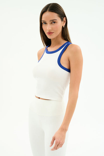 A woman stands against a plain white background wearing the SPLITS59 Kiki Rib Crop Tank in White/Cobalt, featuring soft baby rib fabric, paired with high-waisted white pants.