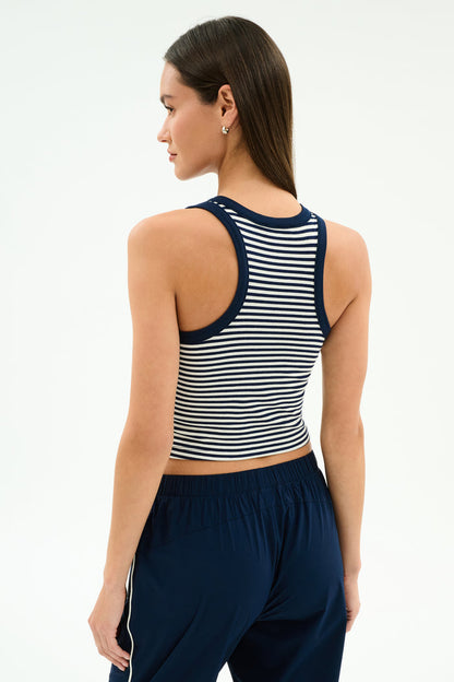 A woman faces away against a plain background, wearing the SPLITS59 Kiki Rib Crop Tank in White/Indigo Stripe with navy pants featuring white side stripes.