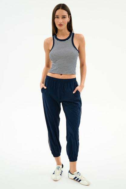 A woman poses with hands in pockets, wearing the SPLITS59 Kiki Rib Crop Tank in White/Indigo Stripe, navy jogger pants, and white Adidas sneakers against a plain white background.