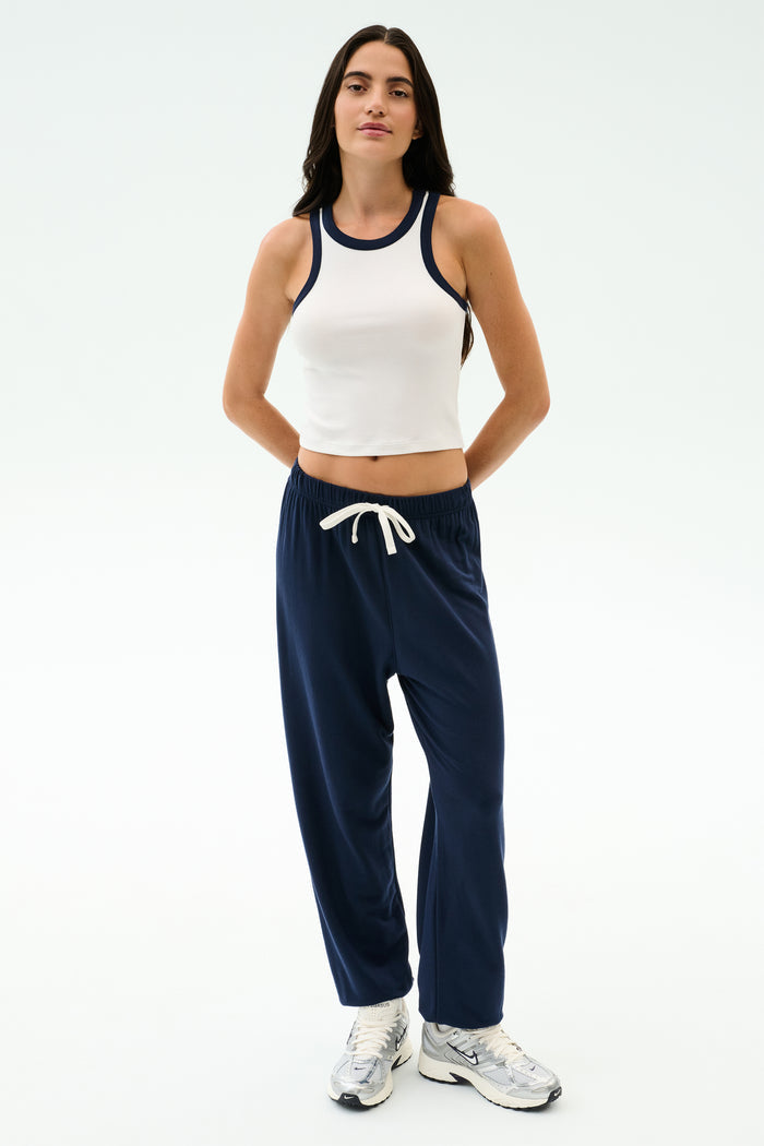 A person stands against a plain background wearing the SPLITS59 Kiki Rib Crop Tank in White/Indigo, navy blue drawstring sweatpants, and white sneakers.