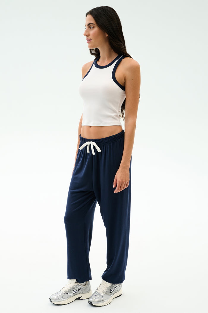 A woman wears the SPLITS59 Kiki Rib Crop Tank in White/Indigo with navy drawstring pants and silver athletic shoes—an easy, gym-ready look against a simple light background.