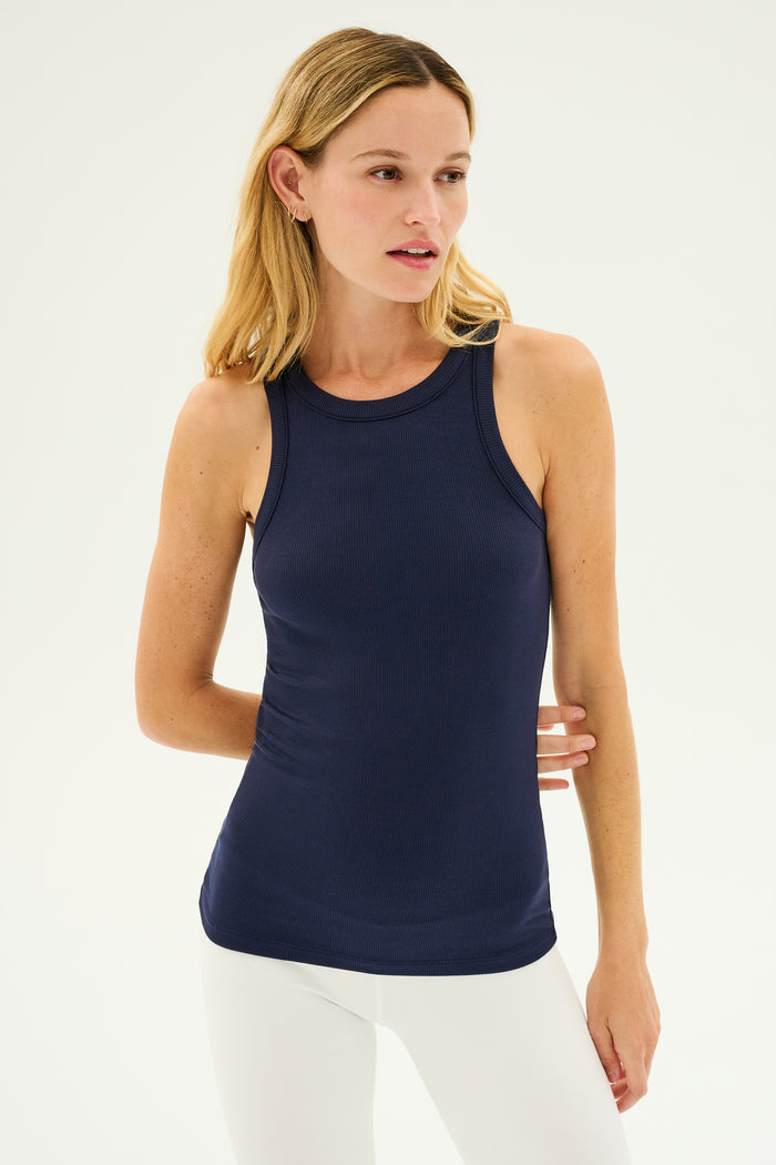 A woman with long blonde hair wears the SPLITS59 Kiki Rib Tank Full Length in Indigo, featuring a high racer neckline, paired with white leggings. She is looking to her right with a neutral expression against a plain background, evoking the essence of dedicated gym workouts.