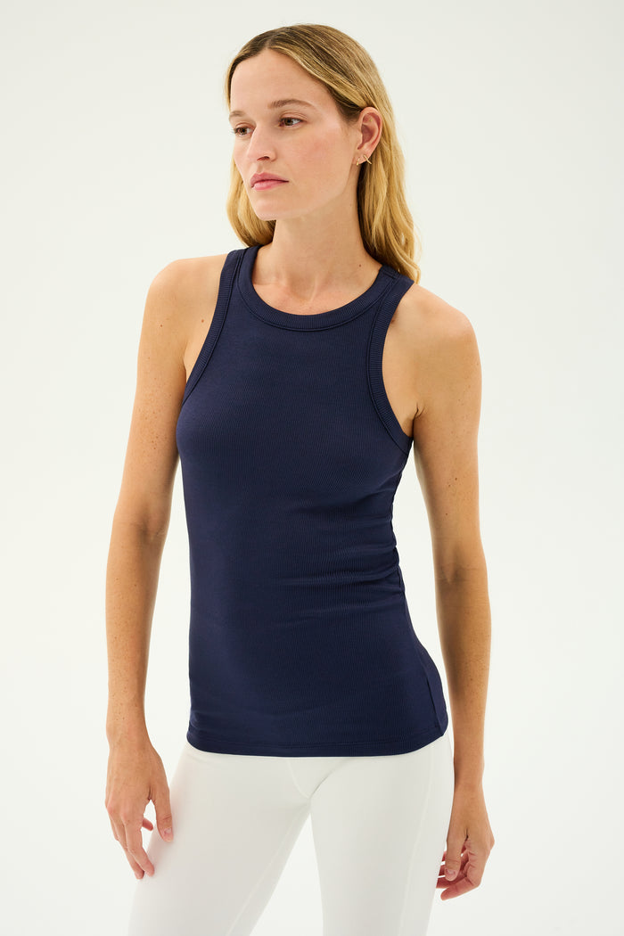 A woman with long blonde hair wearing the SPLITS59 Kiki Rib Tank Full Length in Indigo and white pants stands against a plain background, embodying the elegance of yoga.