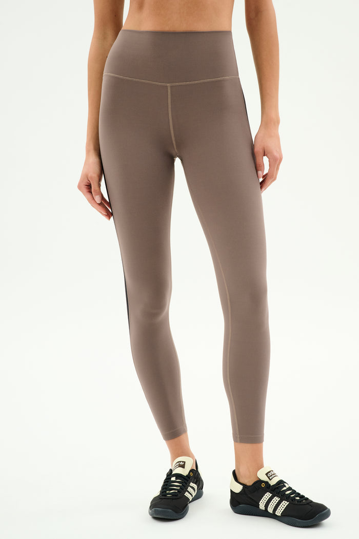 A person wearing SPLITS59 Layla Airweight High Waist 7/8 leggings in Lentil/Black and black sneakers stands against a plain white background—ideal for hot yoga sessions.