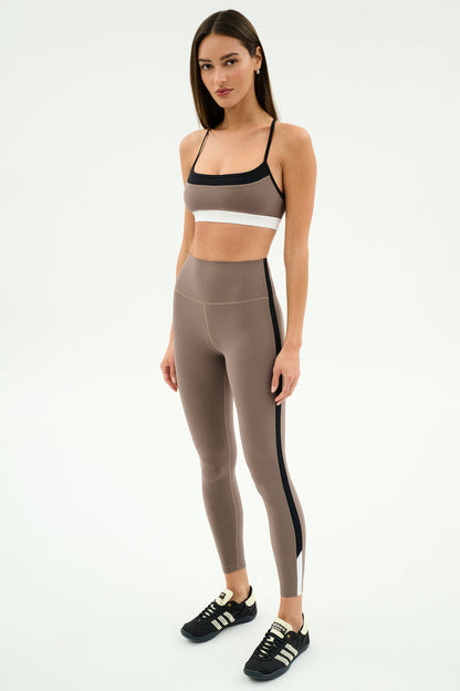 A woman stands against a white background wearing SPLITS59 Layla Airweight High Waist 7/8 leggings in Lentil/Black stripes, paired with a beige sports bra and black athletic shoes—ideal for hot yoga or any workout.