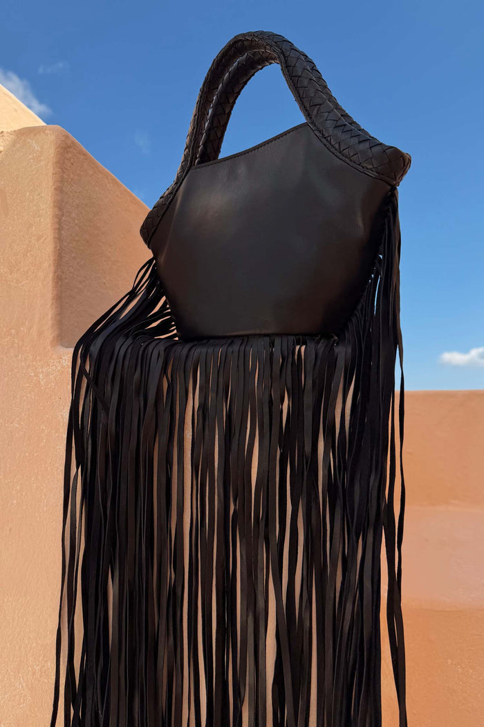Black Leather Fringe Bag featuring a plaited handle and long fringe, displayed outdoors against a blue sky.