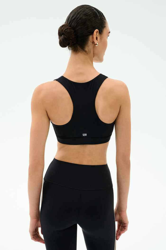 A woman with dark hair in a bun is seen from behind, wearing the SPLITS59 Lexi Rigor Bra in Black/Dark Chocolate and matching leggings—an ideal gym outfit—set against a simple light background.
