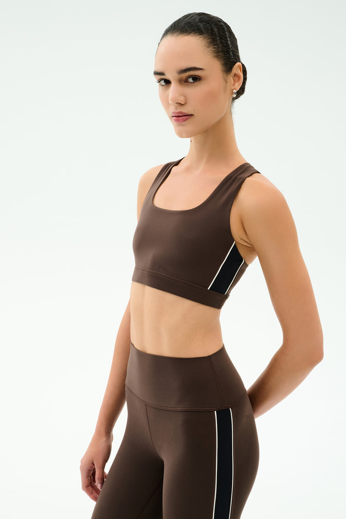 A woman models the SPLITS59 Lexi Rigor Bra in Dark Chocolate/Black, a sports bra-style crop top ideal for gym workouts, paired with matching high-waisted leggings featuring black and white side stripes against a plain light background.