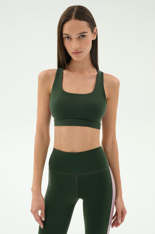 A woman faces forward, wearing the SPLITS59 Lexi Rigor Bra in Military/Blush and matching high-waisted leggings with a white side stripe, set against a plain light background.