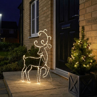 A lighted deer outline holiday decoration standing on a stone doorstep at dusk, with a lit Christmas tree and a brick house in the background.