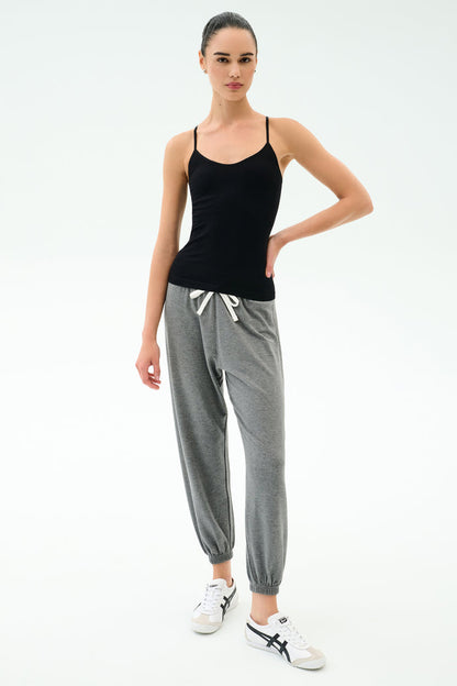 A woman stands against a white background wearing the SPLITS59 Loren Seamless Waist Length Tank in black, paired with gray drawstring jogger pants and white sneakers.