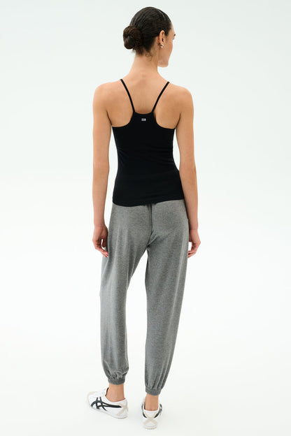 A woman faces away, wearing the SPLITS59 Loren Seamless Waist Length Tank in black, paired with gray jogger pants and white sneakers against a plain light background.
