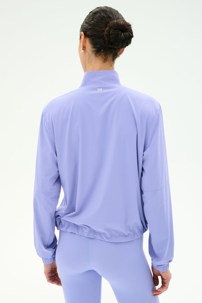A person with dark hair in a bun is seen from the back, wearing the SPLITS59 Lyle Woven Nylon Pullover in Purple Haze/Creme, paired with matching leggings, against a plain white background.