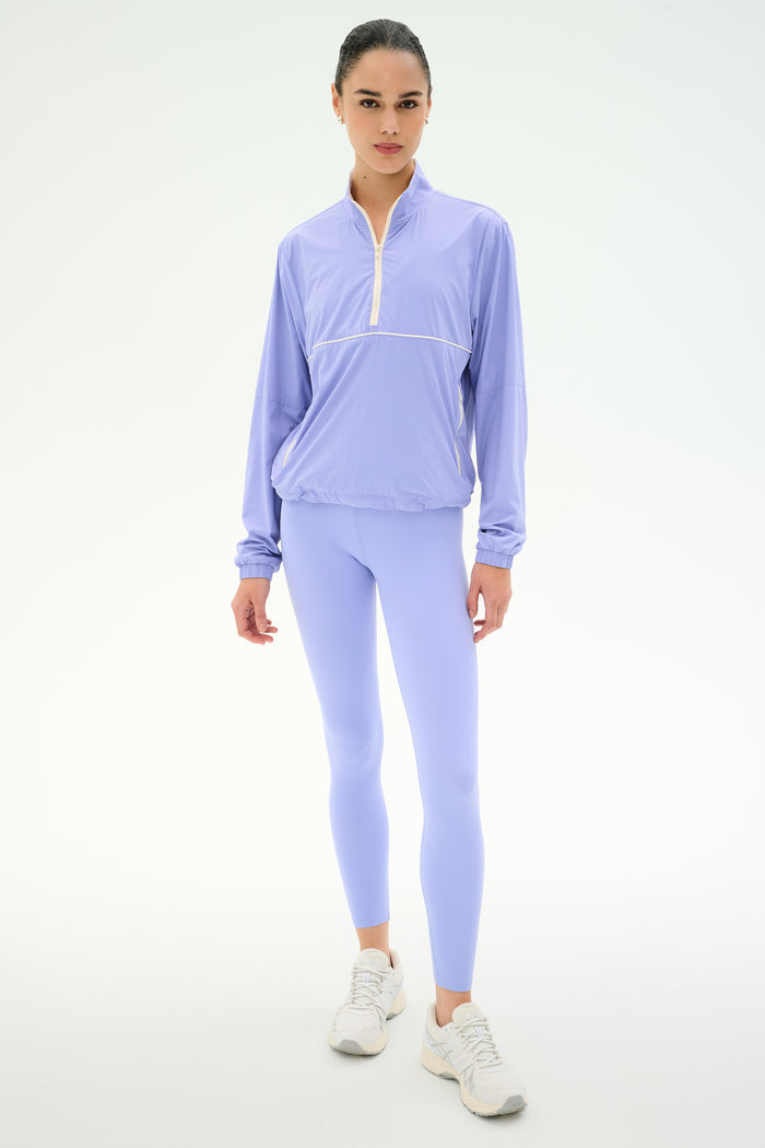 Someone stands facing forward against a white background, wearing the SPLITS59 Lyle Woven Nylon Pullover in Purple Haze/Creme, paired with matching spandex leggings and white sneakers.