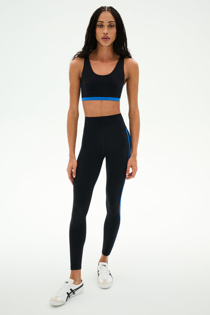 A woman wearing the SPLITS59 Maci Airweight Bra in Black/Classic Blue with matching black leggings featuring blue stripes, paired with white sneakers, stands against a plain white background.