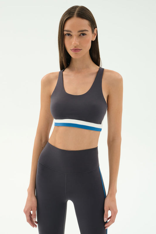 A woman faces forward wearing the SPLITS59 Maci Airweight Bra in Graphite/White with a white and blue band, paired with matching high-waisted leggings, perfect for gym workouts, against a plain light background.