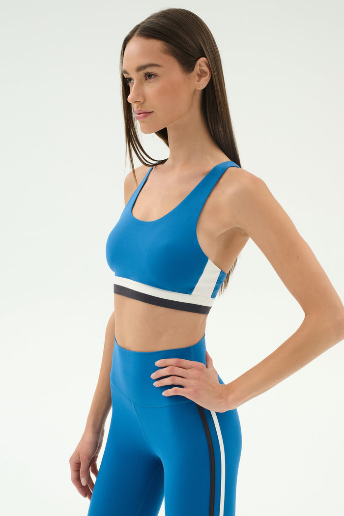 A woman wearing the SPLITS59 Maci Airweight Bra in Stone Blue/Graphite and matching high-waisted leggings with black and white stripes stands with one hand on her hip against a plain background.