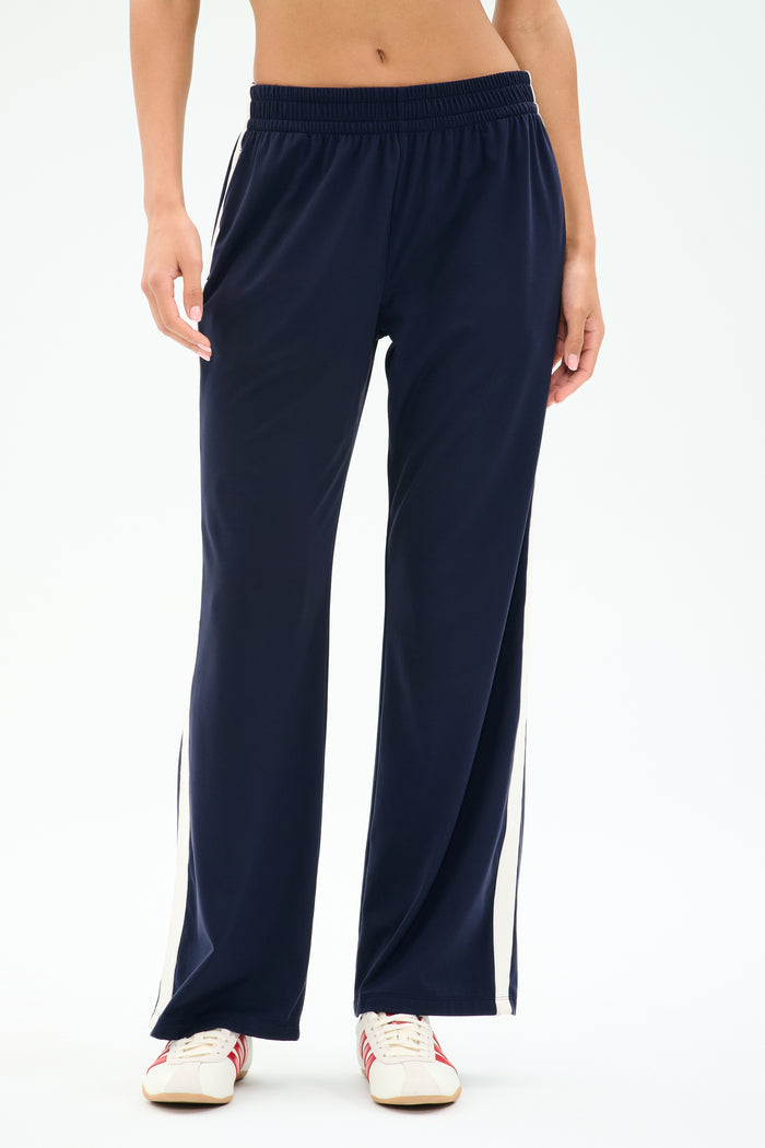 Model wears SPLITS59 Maxie Rigor Track Pant in Indigo/White—navy rigor fabric with white side stripes—paired with white sneakers, shown waist to feet against a plain background.